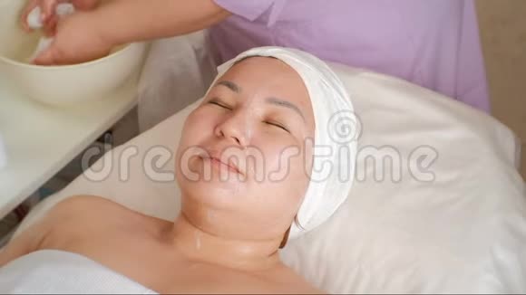 化妆程序一个亚洲女人的特写脸美容师用湿巾擦一个东方女孩的脸和脖子Reju视频的预览图
