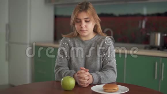 有思想的胖乎乎的女人轮流看厨房里的汉堡和绿苹果的肖像困难的选择视频的预览图