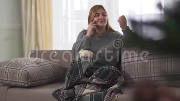 胖乎乎的女人一边微笑一边在智能手机上说话一边坐在沙发上的毯子上加号女性休息视频的预览图
