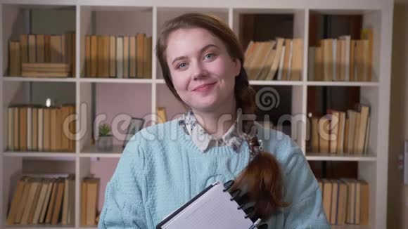一位年轻漂亮的女学生拿着一本书微笑着看着大学图书馆里的照相机的特写照片视频的预览图