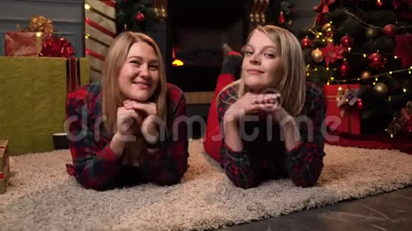 两个穿着红色格子衬衫的漂亮金发女郎躺在靠近新年树或圣诞树的地毯上看着相机视频的预览图