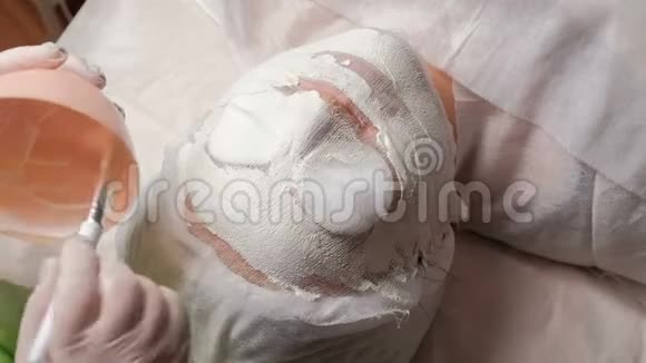 一个美容师或一个戴着白手套的整形外科医生用刷子把一个白色的石膏团涂在女孩脸上纳罗视频的预览图