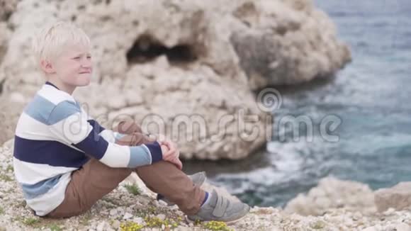 美丽的金发男孩坐在悬崖边的岩石上视频的预览图