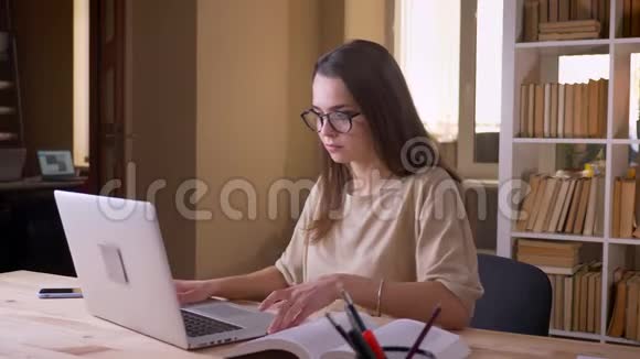 利用笔记本电脑和在大学室内阅读一本书的年轻有魅力的高加索女学生的特写肖像视频的预览图
