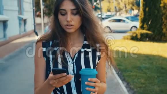 在市中心散步时年轻漂亮的女士可以在前面查看智能手机内容和喝咖啡视频的预览图