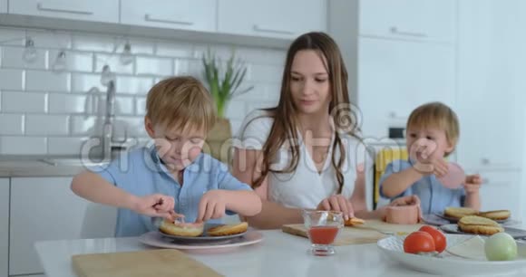 两个弟弟和他们的母亲穿着连衣裙一起在白色的厨房里做饭汉堡笑着笑着视频的预览图