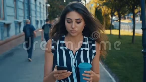 在市中心散步时年轻漂亮的女士可以在前面查看智能手机内容和喝咖啡视频的预览图