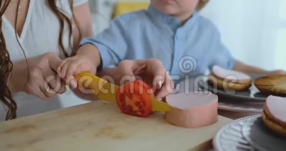 一位年轻的母亲和一个小孩子一起用刀切一个西红柿做自制汉堡健康食品一起烹饪视频的预览图