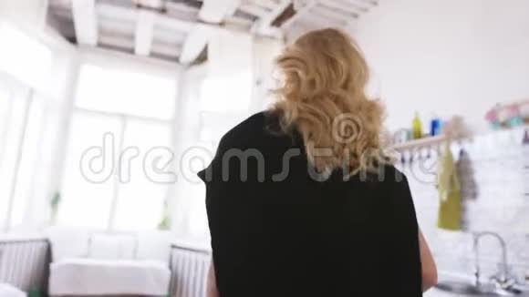穿着白色t恤牛仔裤和黑色套头衫的金发美女在公寓里朝窗户走去视频的预览图