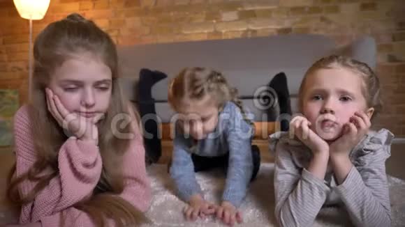 三个小白种人女孩躺在地板上在舒适的家庭氛围中平静地观看镜头的肖像视频的预览图