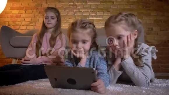 三个白人小女孩躺在地板上在舒适的家庭氛围中严肃地看着平板电脑的画像视频的预览图