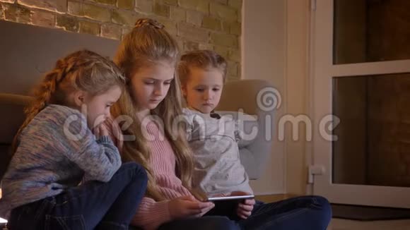 三个漂亮的白种人女孩坐在地板上在舒适的环境中专心地看着平板电脑的画像视频的预览图