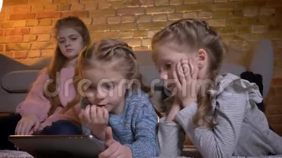 三个小白种人女孩躺在地板上在舒适的家庭氛围中专注地看着平板电脑的肖像视频的预览图