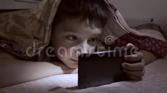 一个男孩躺在家里的床上在毯子下面的黑暗中放松微笑和看智能手机上的视频视频的预览图