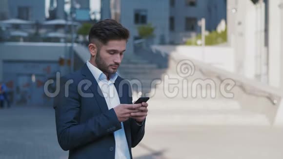 年轻英俊的商人站在大办公楼附近在他的智能手机上输入信息穿着视频的预览图