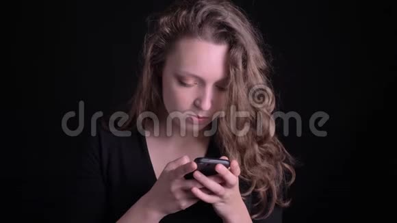 年轻卷发女孩在黑色背景下认真而专注地观看智能手机的肖像视频的预览图