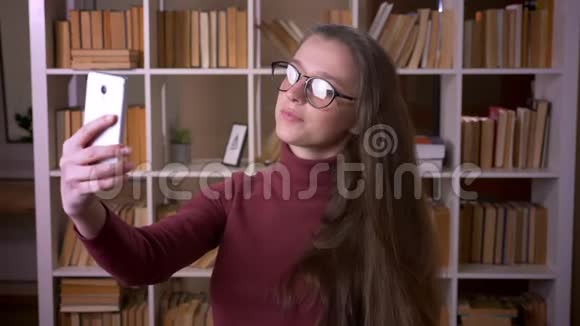戴眼镜的年轻白种人女学生在大学图书馆室内通过手机自拍的特写照片视频的预览图