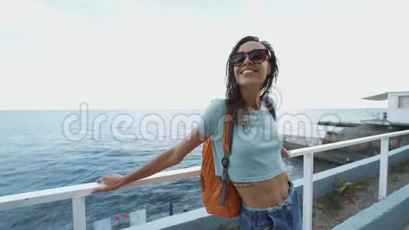 外面的肖像美丽的年轻开朗的微笑女人穿着休闲装和小背包站在海边视频的预览图