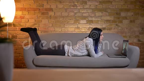 一位年轻漂亮的白种人女性染着头发躺在沙发上用笔记本电脑看电影视频的预览图