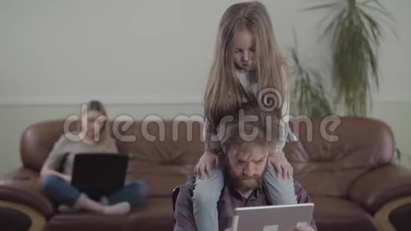有胡子的人用平板电脑特写的肖像悲伤的小女孩坐在父亲的脖子上沙发上的妈妈视频的预览图