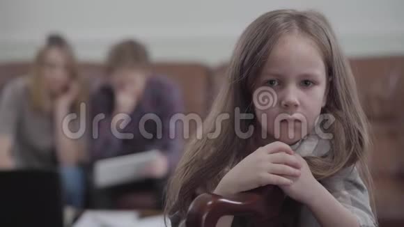 肖像悲伤的小女孩在椅子上看着镜头哭泣年轻女子和长胡子男子的模糊形象视频的预览图