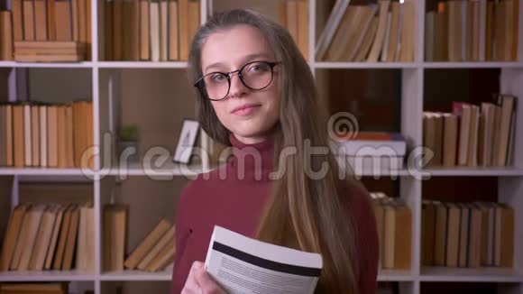 戴着眼镜的年轻白种人女学生的特写照片她拿着一本书开心地笑着看着镜头视频的预览图