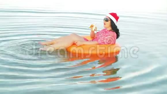 一个游泳圈里的黑发女人和一顶圣诞老人的帽子在海上吃香蕉庆祝圣诞节和新年视频的预览图