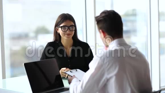 办公室工作人员坐在办公桌前使用笔记本电脑而女性则在后台使用电话视频的预览图