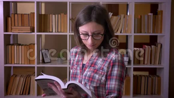 一位戴眼镜的年轻有魅力女学生的特写镜头她在图书馆看书微笑着看着摄像机视频的预览图