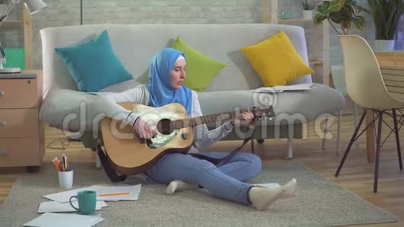 年轻的穆斯林妇女戴着头巾坐在地板上弹吉他和录音和弦视频的预览图