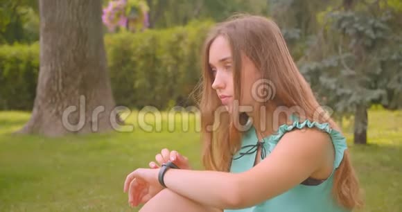 在户外公园里年轻可爱的长发美丽的女性坐在绿草上用智能手表特写肖像视频的预览图