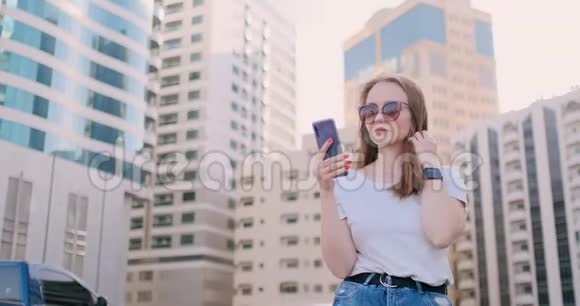 年轻的美女手拿智能手机对着夏天的摩天大楼微笑视频的预览图