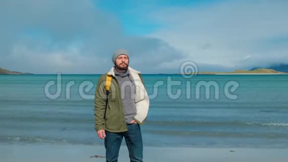 穿着绿色夹克和黄色背包的旅行者站在海滩上站在高山峰的背景下视频的预览图