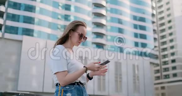 年轻的美女手拿智能手机对着夏天的摩天大楼微笑视频的预览图