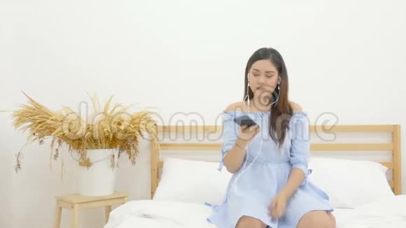 4K美丽的亚洲女人坐在床上用手机上的耳机听音乐跳舞视频的预览图
