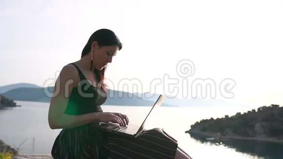 一个漂亮的女孩在一台笔记本电脑上工作坐在海面上的岩石上慢动作视频的预览图