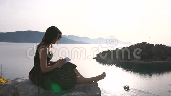 一个迷人的女孩坐在高高的海面上的岩石上写着一本笔记本视频的预览图