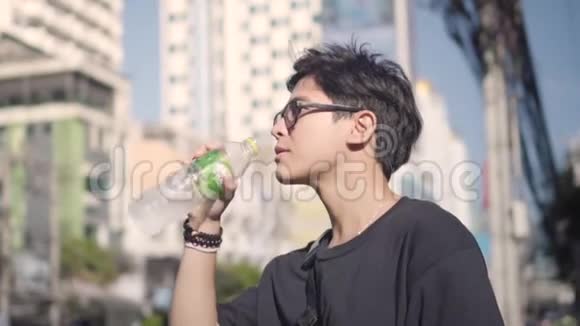 亚洲男人在炎热的日子里从瓶子里喝水背景中的城市景观视频的预览图