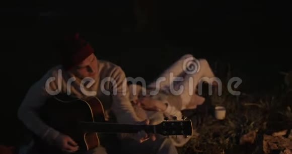 一对年轻夫妇晚上坐在火边这家伙弹吉他他的女朋友躺在他的腿上听着视频的预览图