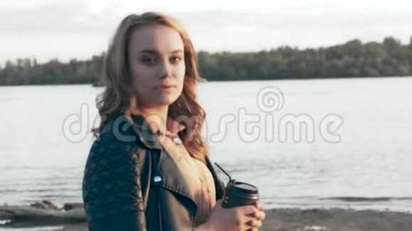 穿着黑色夹克的漂亮女孩站在河边喝咖啡视频的预览图