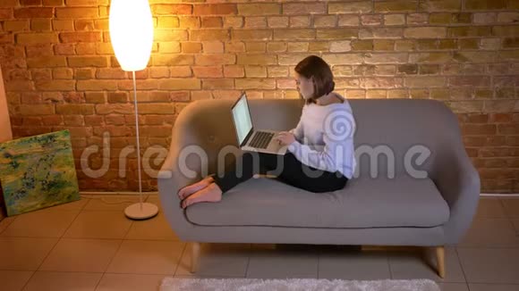 年轻的高加索女性在笔记本电脑上打字坐在沙发上做远程工作的特写镜头视频的预览图