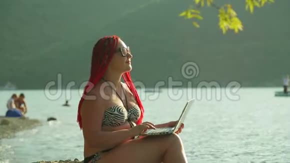 一个红头发的女人在岸边贴着群山的背景写博客视频的预览图