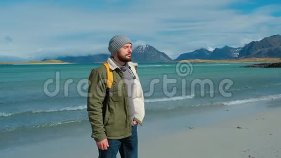 穿着绿色夹克和黄色背包的旅行者站在海滩上站在高山峰的背景下视频的预览图