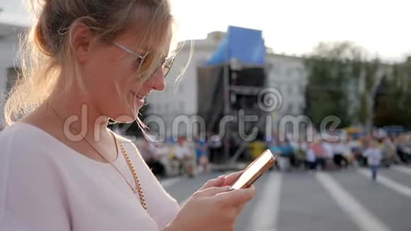 戴眼镜的可爱女人手里拿着智能手机在市中心的阳光下微笑视频的预览图