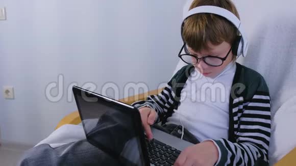 男孩用耳机和坐在扶手椅上的眼镜在笔记本电脑上写和键入信息视频的预览图