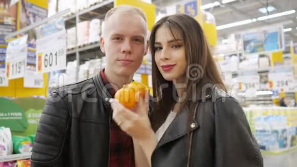 在超市的年轻夫妇女孩在做滑稽的鬼脸橘子在超市里看着相机视频的预览图