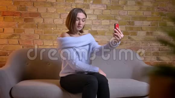 一位年轻迷人的白种人女性在舒适的家中坐在沙发上用电话自拍的特写镜头视频的预览图