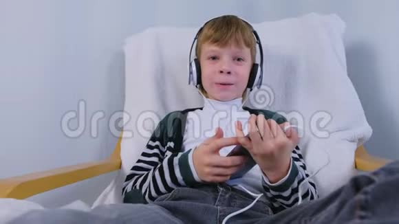 男孩在智能手机里用耳机听音乐坐在家里的扶手椅上唱歌视频的预览图