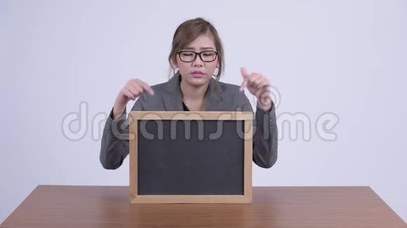 年轻严肃的亚洲女商人在书桌前展示黑板和大拇指视频的预览图