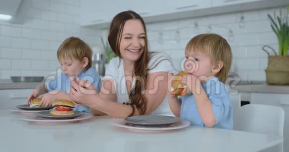 小男孩坐在厨房里他的母亲和弟弟吃着汉堡微笑着健康食品家庭汉堡视频的预览图